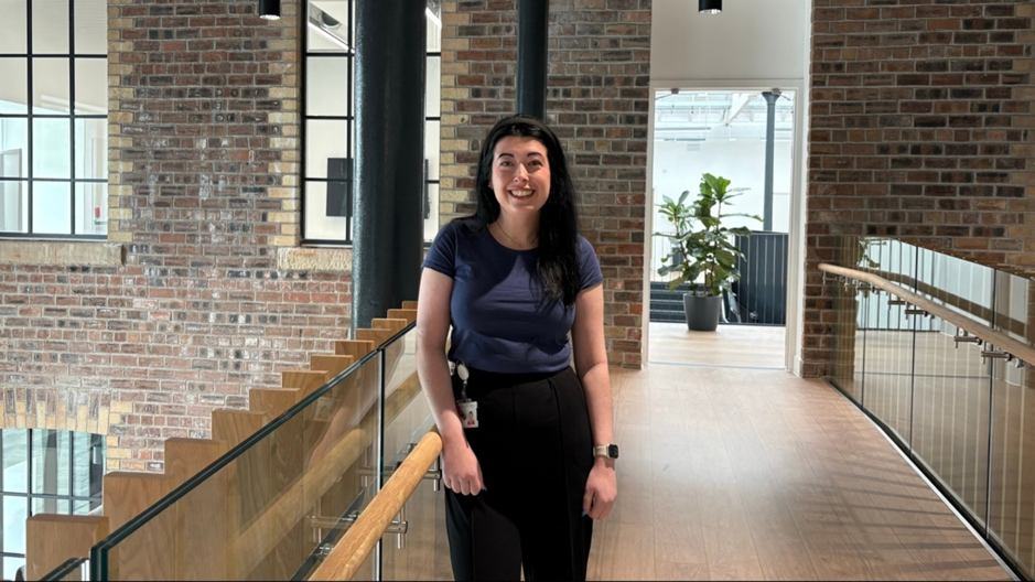Person standing on an indoor balcony walkway in a modern building with exposed brick walls and large windows. The person is positioned beside a wooden handrail and glass barrier, wearing a dark top and trousers, with a staff ID badge visible. In the background are additional walkways, a staircase, and a potted plant in a bright open space.
