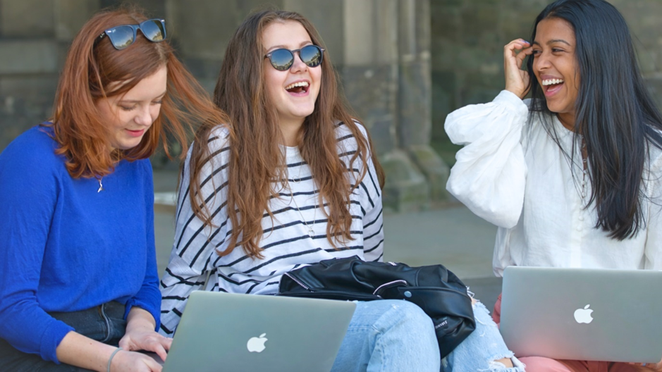 Three people sitting outdoors on steps with open laptops, laughing and chatting together. One person in a blue top looks down at a laptop, another in a striped top wears sunglasses and sits with a bag on their lap, and a third in a white top holds a laptop while adjusting their hair.
