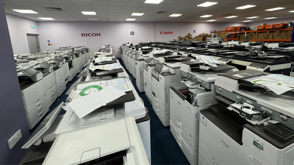A large warehouse room containing rows of multifunction printers and photocopiers arranged closely together. Equipment from brands including Ricoh, HP, and Canon is visible, with company logos displayed on the far wall. Many machines have inspection sheets or paperwork placed on top, suggesting testing, refurbishment, or preparation for resale.