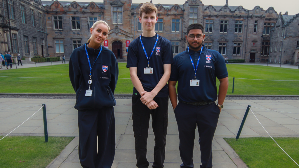Three University of St Andrews IT Services staff members wearing navy branded uniforms, lanyards, and identification badges stand on a paved walkway in a historic university courtyard. A large stone academic building with arched entrances, decorative masonry, and rows of windows forms the backdrop, while a well-maintained lawn stretches across the centre of the courtyard.
